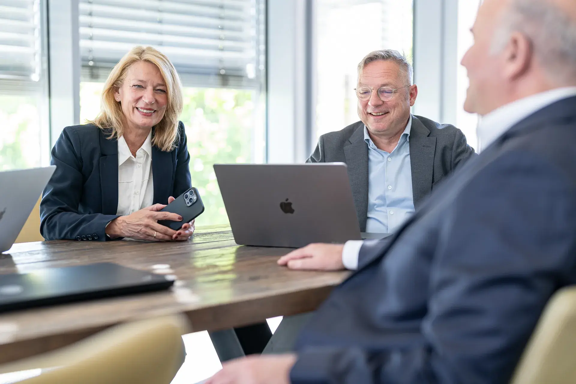 Beraterin und Berater lachen im Gespräch mit einem Kollegen am Holztisch mit Laptops