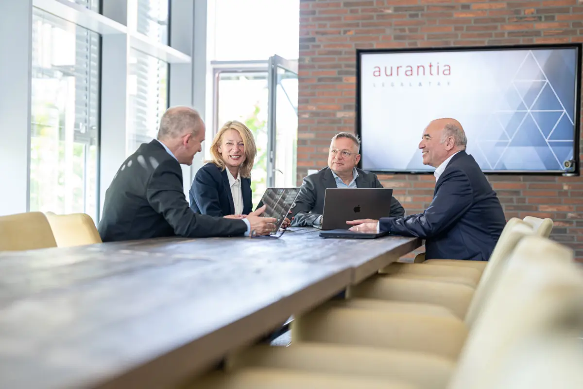 Vier Berater im Gespräch am Konferenztisch, im Hintergrund ein Bildschirm mit aurantia-Logo vor Backsteinwand