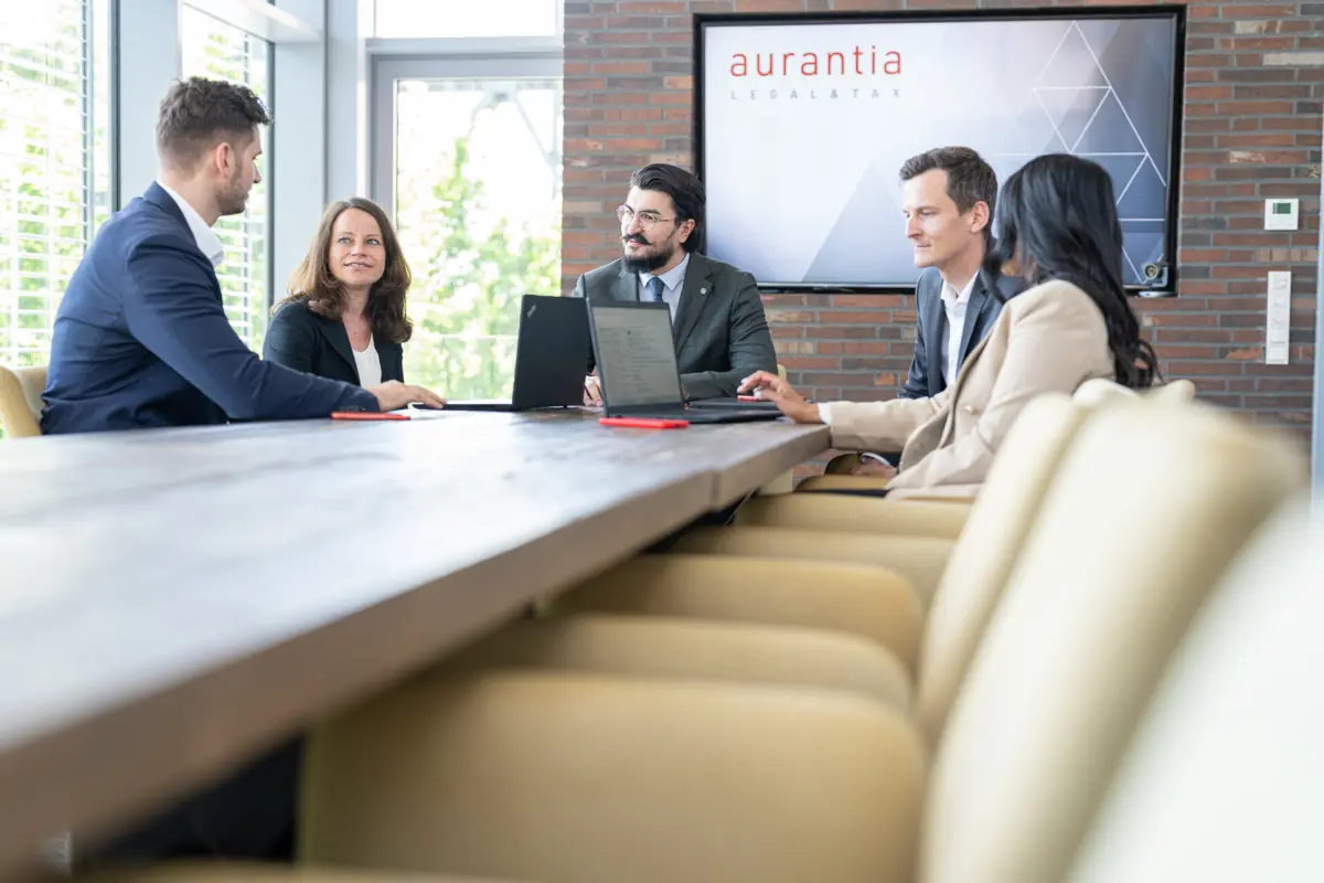 Fünf Beraterinnen und Berater beim Meeting am langen Tisch mit Laptops vor dem aurantia-Bildschirm