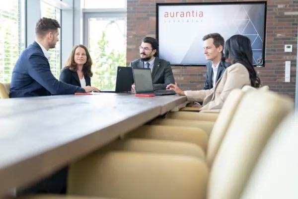 Fünf Beraterinnen und Berater beim Meeting am langen Tisch mit Laptops vor dem aurantia-Bildschirm