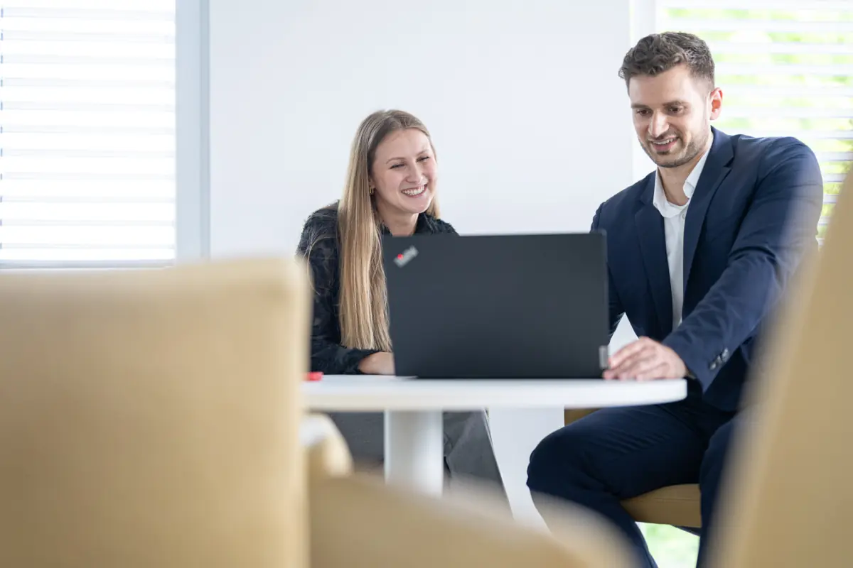 Lachende Beraterin und Berater schauen an einem runden Tisch gemeinsam auf einen Laptop