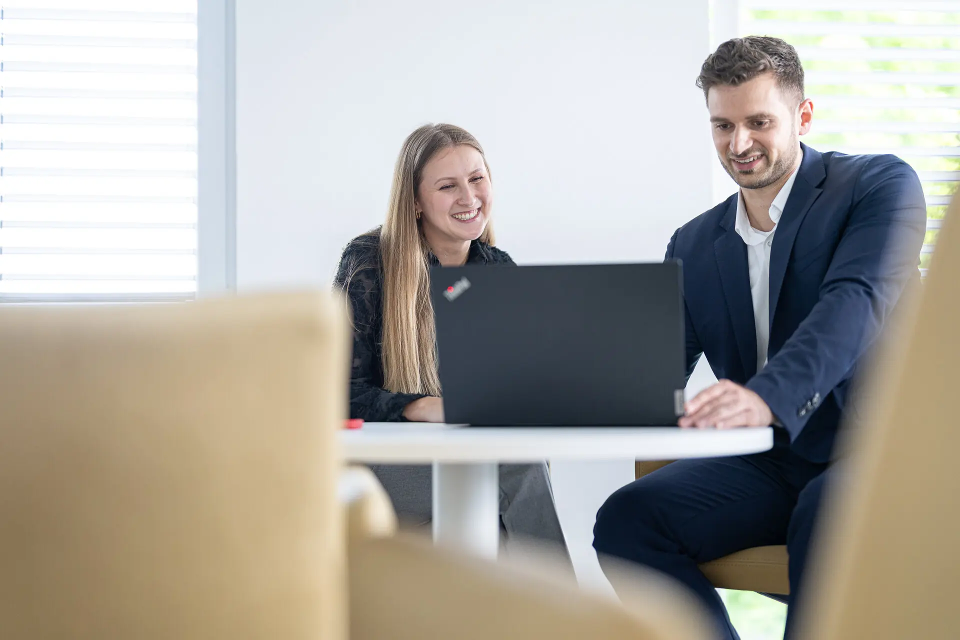 Lachende Beraterin und Berater schauen an einem runden Tisch gemeinsam auf einen Laptop