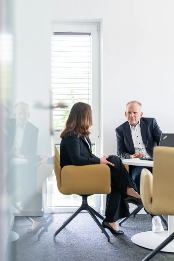 Beraterin und Berater im Beratungsgespräch an einem runden Tisch in einem hellen Büro hinter einer Glasscheibe