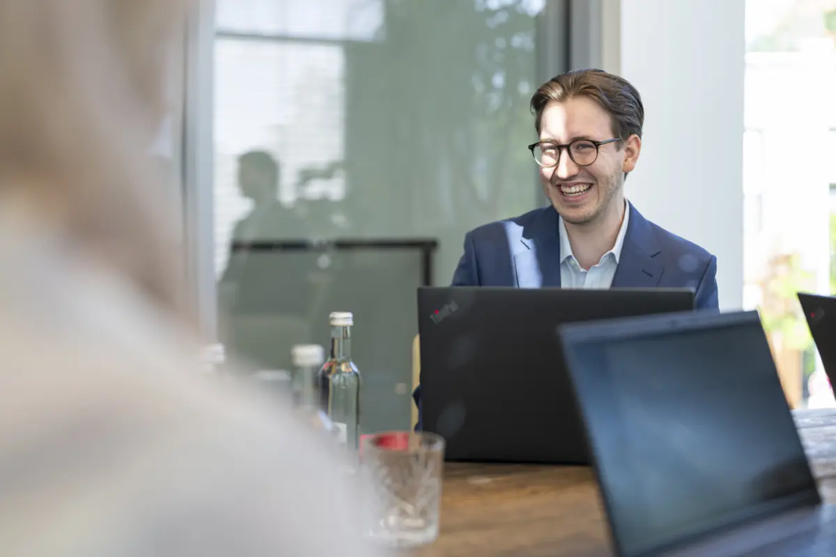 Lachender junger Berater mit Brille und blauem Sakko hinter seinem Laptop am Besprechungstisch