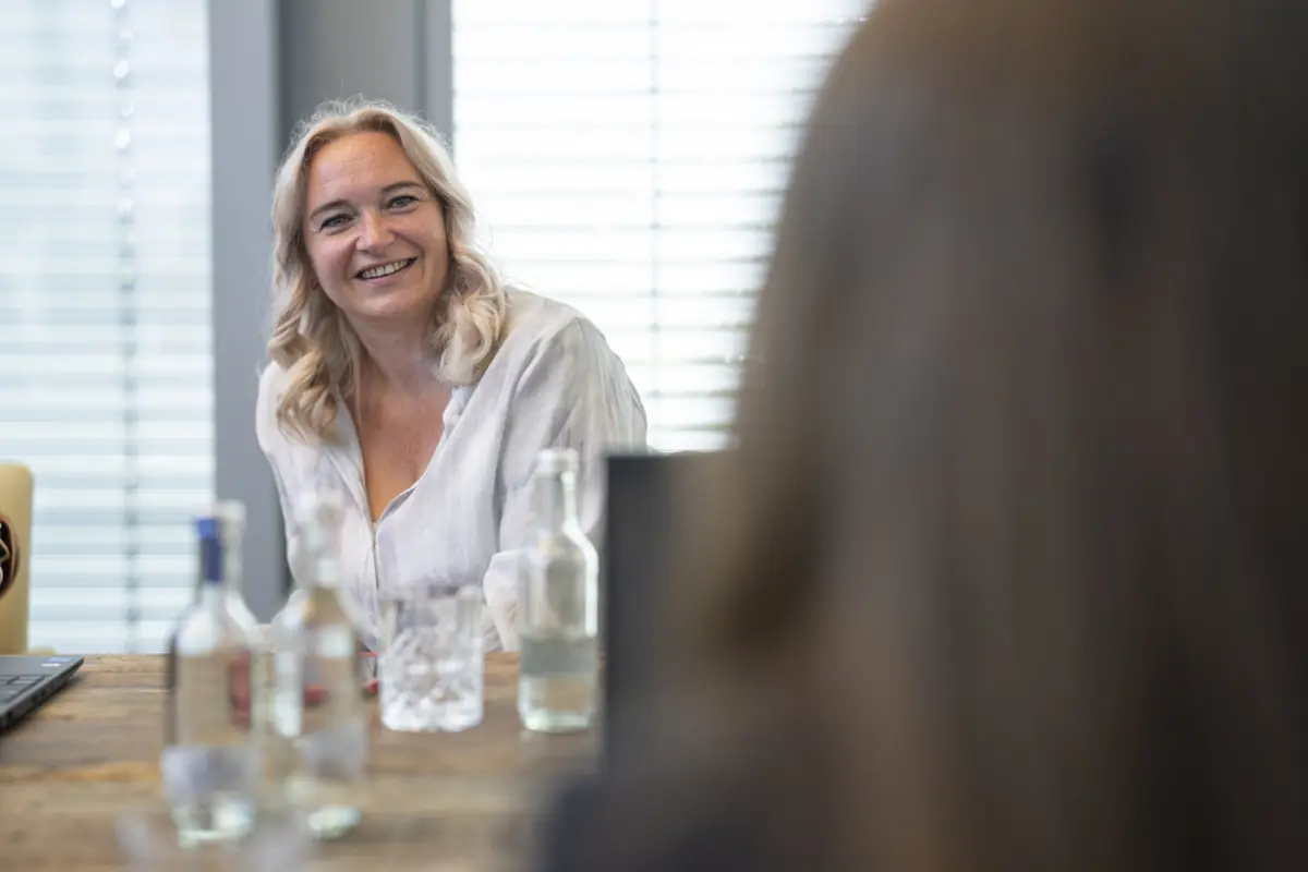 Lächelnde Beraterin mit blonden Haaren am Besprechungstisch, im Vordergrund unscharf ein Gesprächspartner