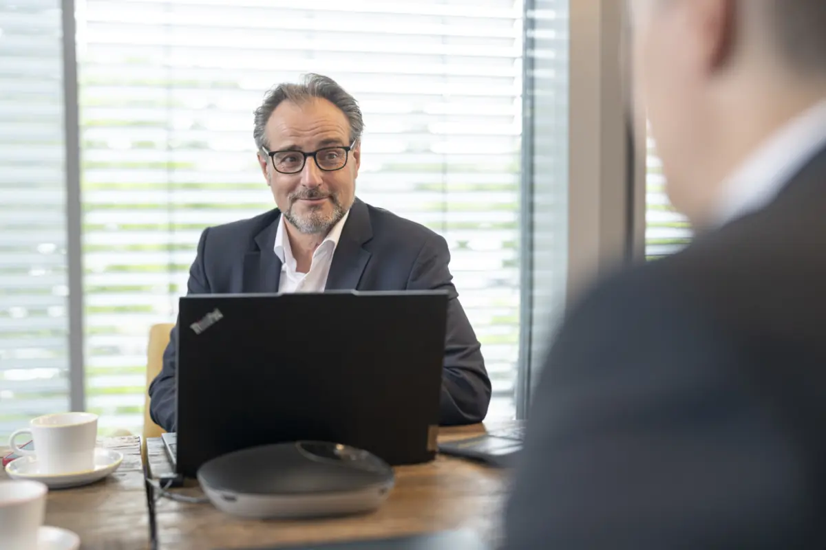 Berater mit Brille sitzt hinter einem Laptop mit Konferenztelefon am Holztisch und blickt zu einem Gesprächspartner