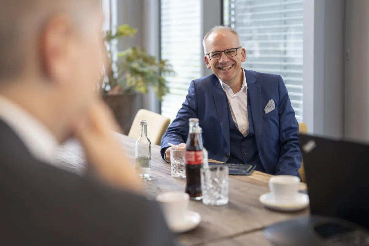 Lachender Berater mit Brille und blauem Anzug am Holztisch mit Getränken, Kaffeetassen und Laptop