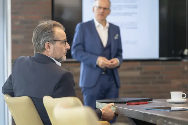 Berater hört bei einer Präsentation zu, im Hintergrund steht ein Kollege vor einem Bildschirm
