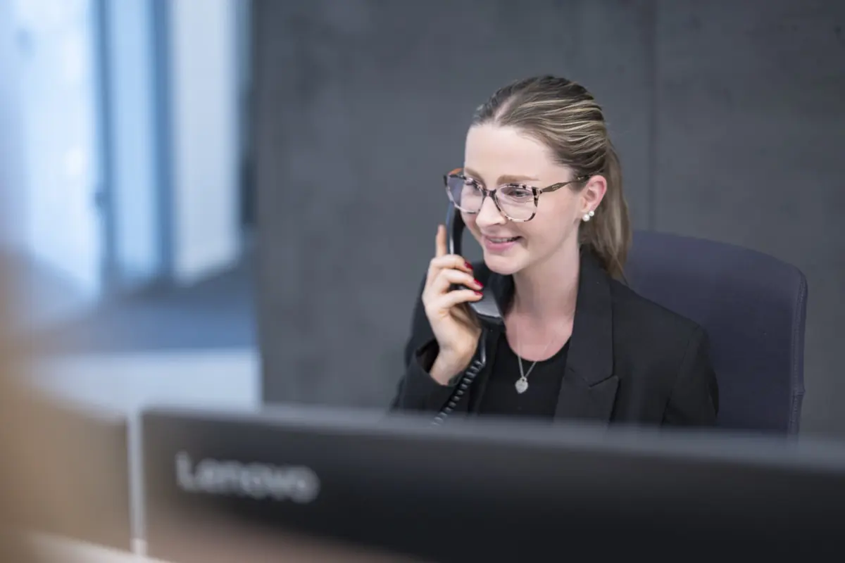Mitarbeiterin mit Brille telefoniert lächelnd am Empfangsplatz vor einem Monitor