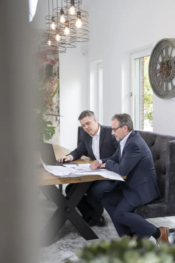 Zwei Berater in dunklen Anzügen besprechen Planunterlagen und Laptop an einem Holztisch in einem hellen Raum
