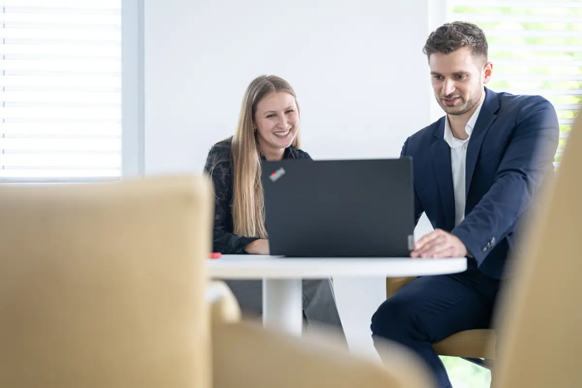 Eine Beraterin und ein Berater sitzen lächelnd an einem runden Tisch und schauen gemeinsam auf einen Laptop