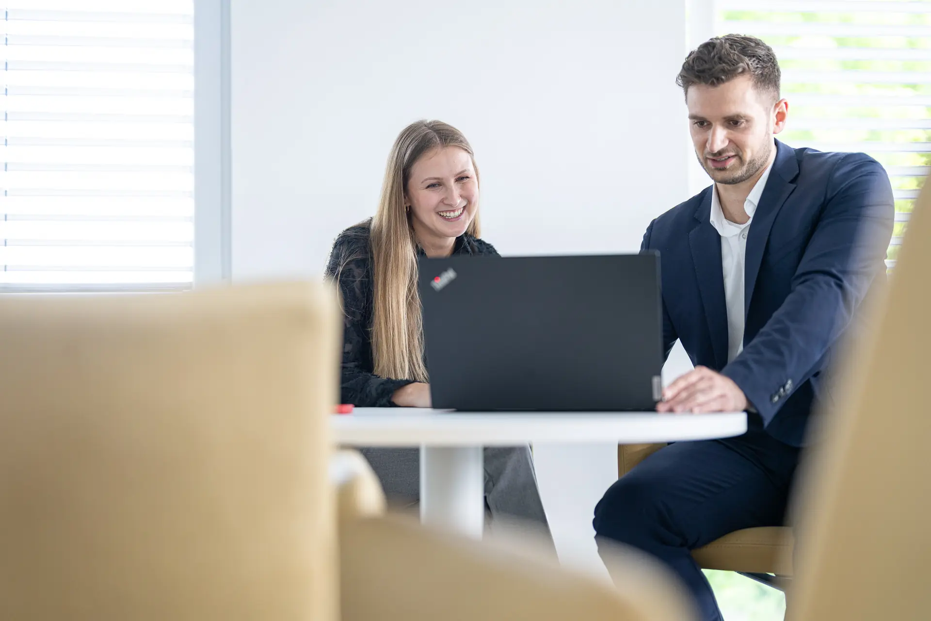 Eine Beraterin und ein Berater sitzen lächelnd an einem runden Tisch und schauen gemeinsam auf einen Laptop