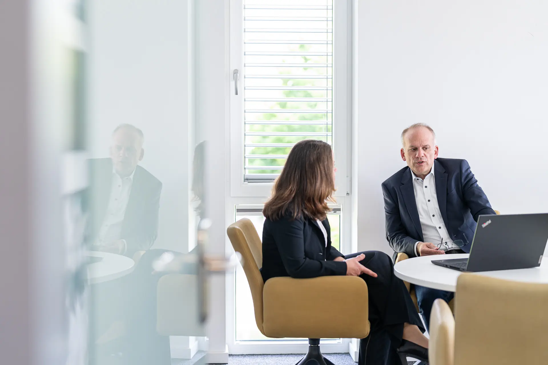 Eine Beraterin und ein Berater im Gespräch an einem runden Tisch mit Laptop in einem hellen Büro