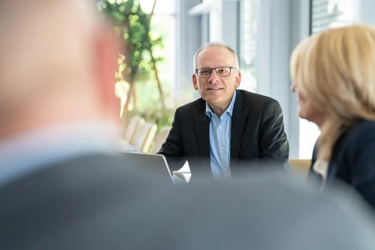 Berater mit Brille blickt im Besprechungsraum freundlich in die Kamera, im Vordergrund zwei unscharfe Kollegen