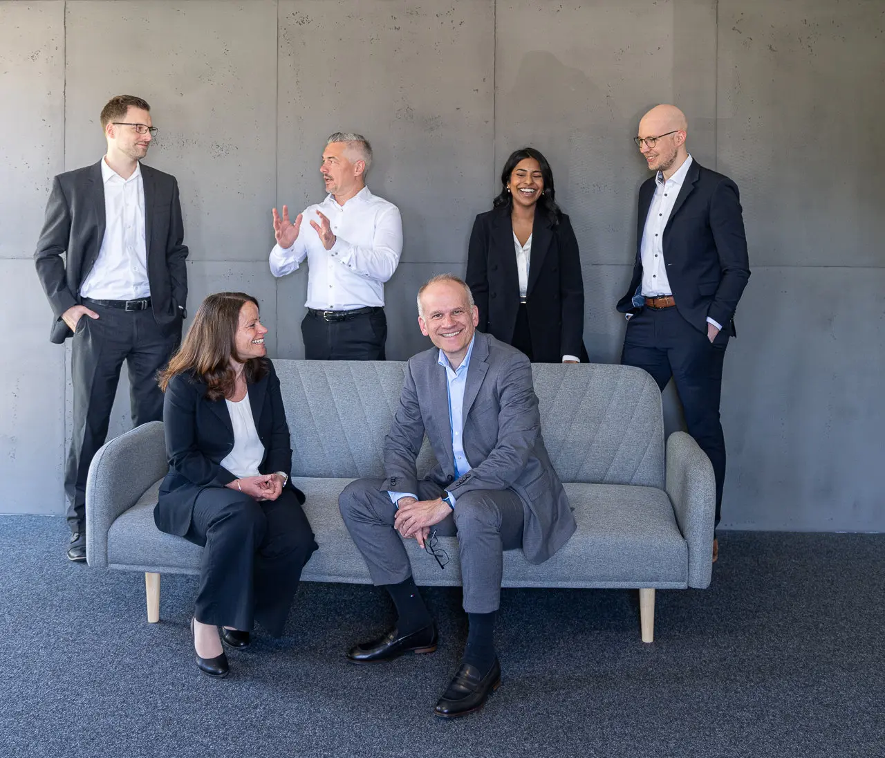 Sechs Berater und Beraterinnen im Business-Outfit lachen und unterhalten sich an einem grauen Sofa vor einer Betonwand
