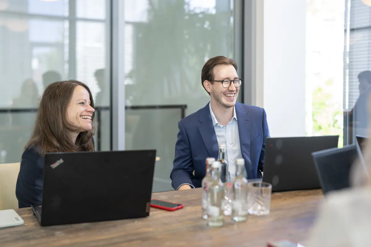 Eine Beraterin und ein Berater lachen bei einer Besprechung am Holztisch, vor ihnen Laptops und Wasserflaschen